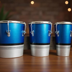 Può includere: Tre conga blu con finiture argentate e base argento e nera sono disposte su una superficie di legno. I tamburi sono di dimensioni variabili, con una lucentezza metallica. Lo sfondo è sfocato, con un'illuminazione calda.
