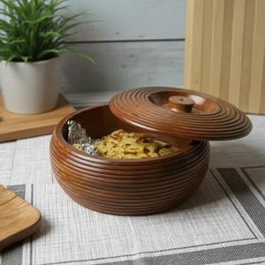 May include: A round, dark brown wooden container with a matching lid, both featuring horizontal ribbed detailing. The container is partially open, revealing a piece of flatbread wrapped in silver foil inside the bowl-shaped base.