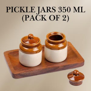 Puede incluir: Dos tarros de encurtidos de cerámica marrón y blanco con tapas, cada uno con una capacidad de 350 ml, sobre una bandeja de madera. El texto "PICKLE JARS 350 ML (PACK OF 2)" se muestra sobre los tarros.