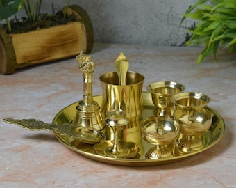 May include: A golden brass puja thali set with various ritual items. The set includes a bell, a cup with a spoon, small bowls, and a decorative spoon, all arranged on a round tray. The items are made of shiny brass.