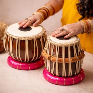 Puede incluir: Dos tambores tabla marrones con parches blancos y detalles tejidos descansan sobre bases circulares rosas con ribetes dorados. Las manos de una persona tocan los tambores, adornadas con anillos y pulseras. Los tambores son un instrumento de percusión indio tradicional.