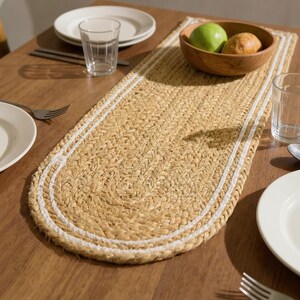 Puede incluir: Un camino de mesa de forma ovalada elaborado con fibras de yute trenzadas en color tostado natural, que presenta un detalle de borde cosido en blanco que sigue el contorno redondeado de la pieza, ofreciendo una superficie texturizada.