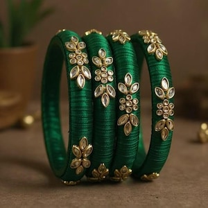 May include: A set of four emerald green bangles wrapped in silk thread. Each bangle features gold-toned metal accents arranged in floral and leaf-like patterns, embedded with clear, reflective stones that catch the light along the outer surface.