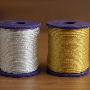 May include: Two cylindrical spools of thread, one wound with white cord and the other with gold cord. Each spool features purple plastic end caps with a circular hole in the center, providing a structured base for the tightly wound metallic-toned thread.