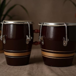 May include: A pair of brown bongo drums with a light brown stripe. The drums have silver hardware and are set against a neutral background. The drums are a musical instrument.