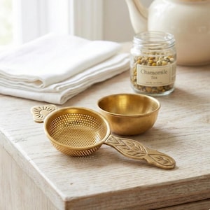 Puede incluir: Un infusor de té dorado con un diseño de sol y un cuenco pequeño sobre una superficie de madera clara. Un tarro de cristal con té de manzanilla y una tetera blanca en el fondo. También se ven sábanas blancas dobladas.