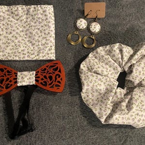 May include: A set of accessories including a red wooden bow tie with a floral design, a white and purple floral patterned handkerchief, a white and purple floral patterned scrunchie, and a pair of gold earrings with white and purple floral patterned accents.