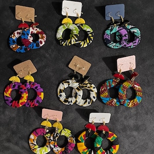 Peut inclure: Un ensemble de neuf paires de boucles d'oreilles en tissu coloré en forme de cercle. Chaque paire présente un tissu imprimé africain différent avec un cercle doré et un accent doré ou rouge. Les boucles d'oreilles sont présentées sur un fond noir.