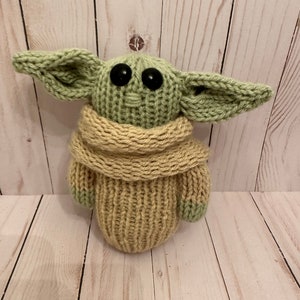 May include: A knitted green and beige plush toy with large black eyes. The toy is wearing a beige knitted scarf.