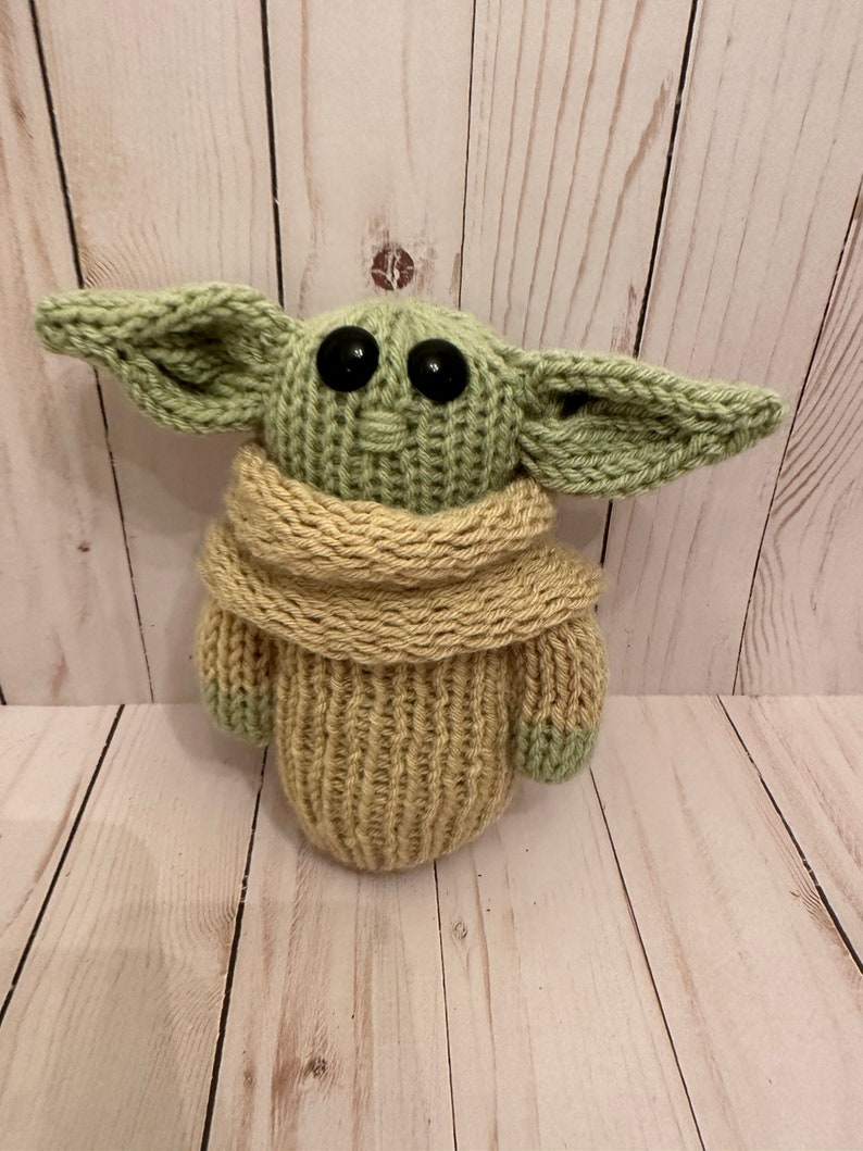 May include: A knitted green and beige plush toy with large black eyes. The toy is wearing a beige knitted scarf.