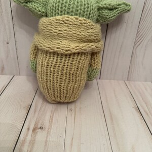 May include: A knitted green and tan plush toy resembling a character from the Star Wars franchise. The toy is wearing a tan knitted coat.