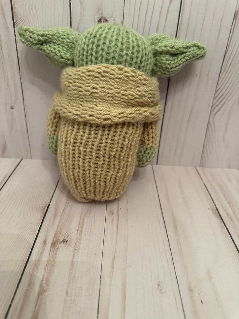 May include: A knitted green and tan plush toy resembling a character from the Star Wars franchise. The toy is wearing a tan knitted coat.
