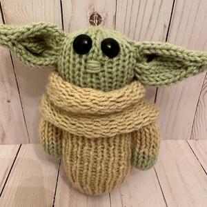 May include: A knitted green and beige plush toy resembling a character from the Star Wars franchise. The toy has large black eyes and a beige scarf.
