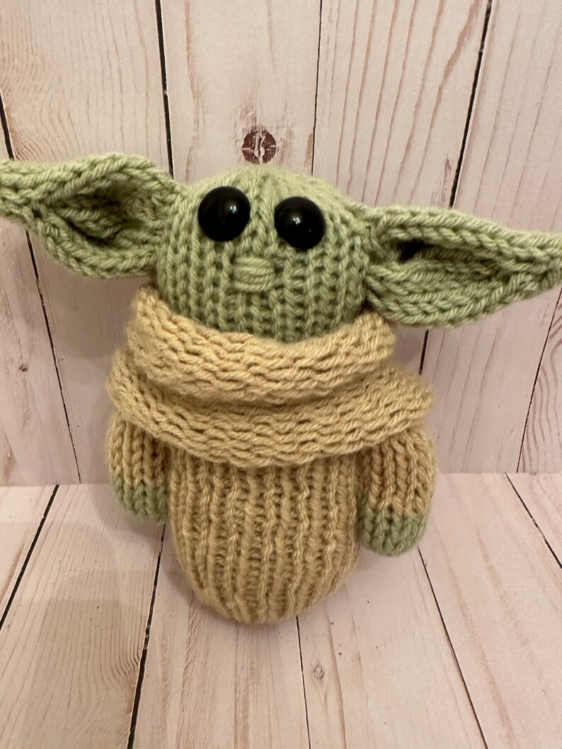 May include: A knitted green and beige plush toy resembling a character from the Star Wars franchise. The toy has large black eyes and a beige scarf.