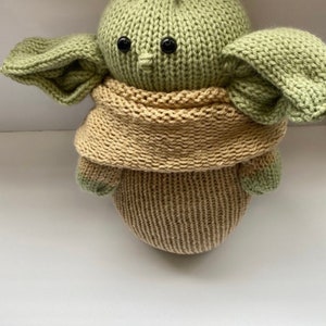 May include: A knitted green and beige plush toy with large ears and black eyes. The toy is wearing a beige knitted scarf.