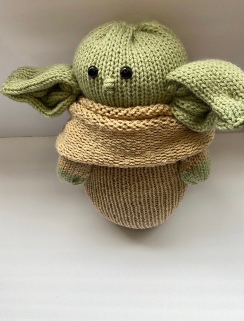 May include: A knitted green and beige plush toy with large ears and black eyes. The toy is wearing a beige knitted scarf.