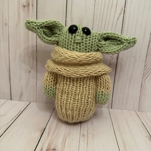 May include: A knitted green and tan plush toy resembling a character from the Star Wars franchise. The toy has large ears, black eyes, and a tan knitted sweater.