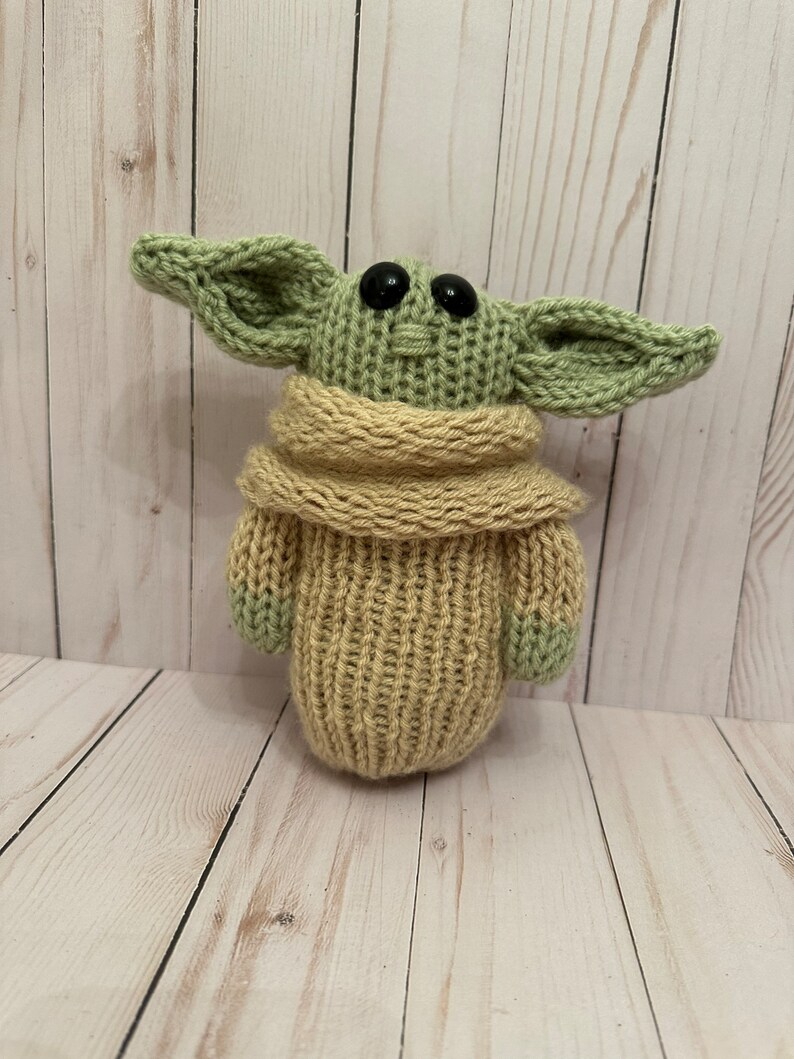 May include: A knitted green and tan plush toy resembling a character from the Star Wars franchise. The toy has large ears, black eyes, and a tan knitted sweater.