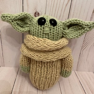 May include: A knitted green and beige plush toy with large black eyes. The toy is wearing a beige knitted scarf.