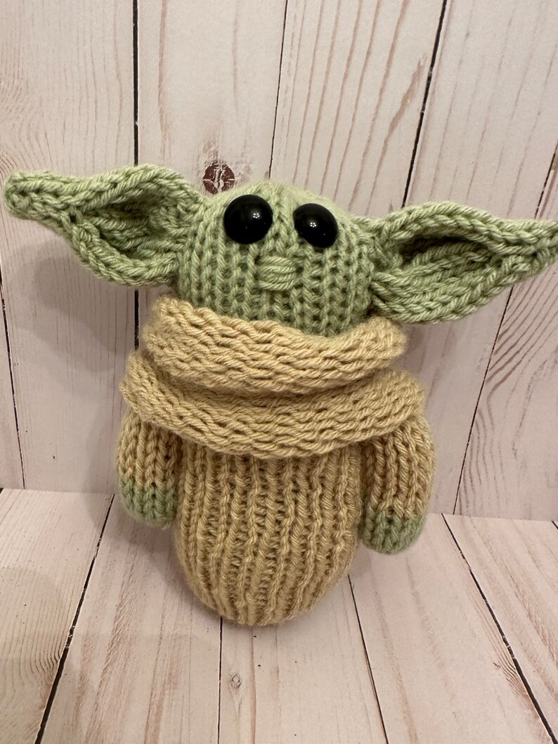 May include: A knitted green and beige plush toy with large black eyes. The toy is wearing a beige knitted scarf.