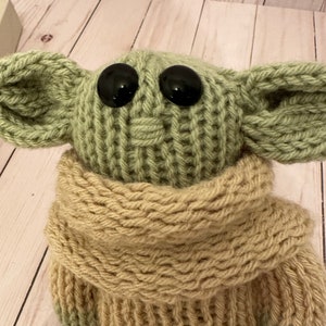 May include: A knitted green and beige plush toy with large black eyes. The toy is wearing a beige knitted scarf.