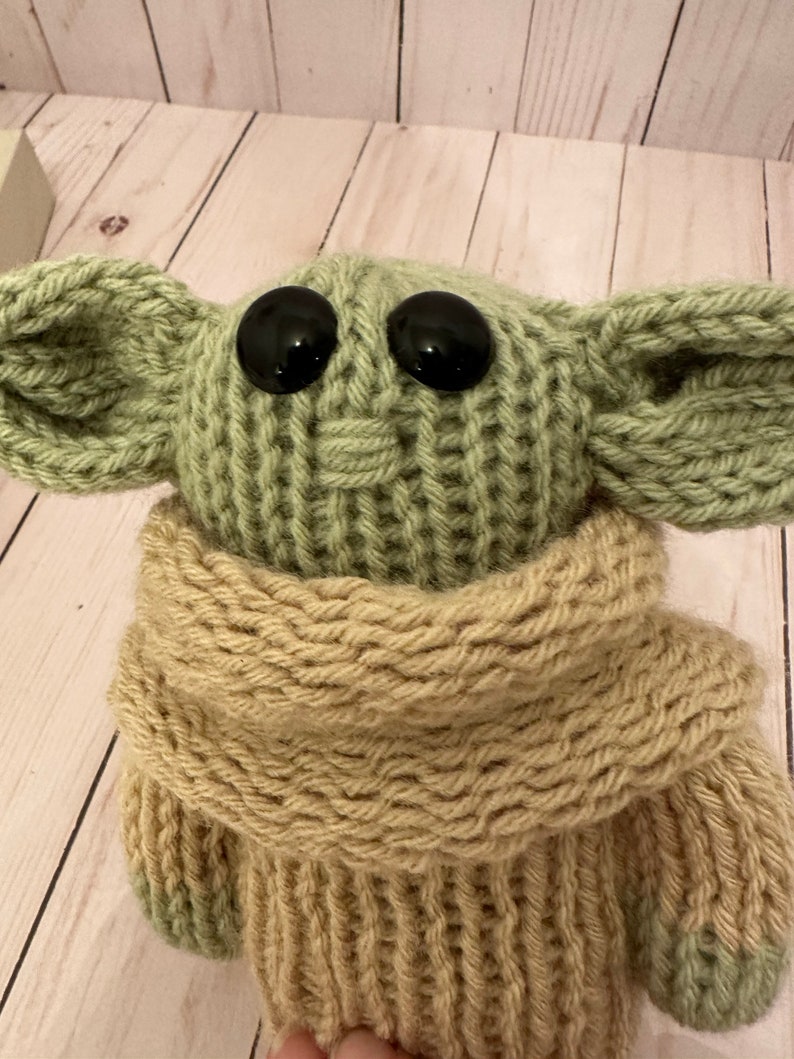 May include: A knitted green and beige plush toy with large black eyes. The toy is wearing a beige knitted scarf.