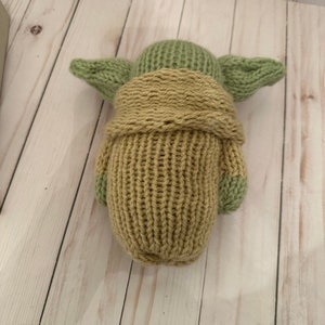 May include: A knitted plush toy of a green character with large ears, wearing a brown robe. The toy is made with a soft, textured yarn.