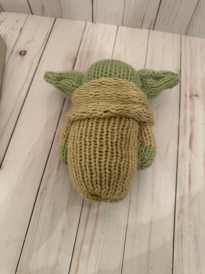 May include: A knitted plush toy of a green character with large ears, wearing a brown robe. The toy is made with a soft, textured yarn.