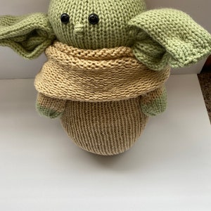 May include: A knitted green and tan plush toy resembling a popular fictional character. The toy has large ears and a scarf.