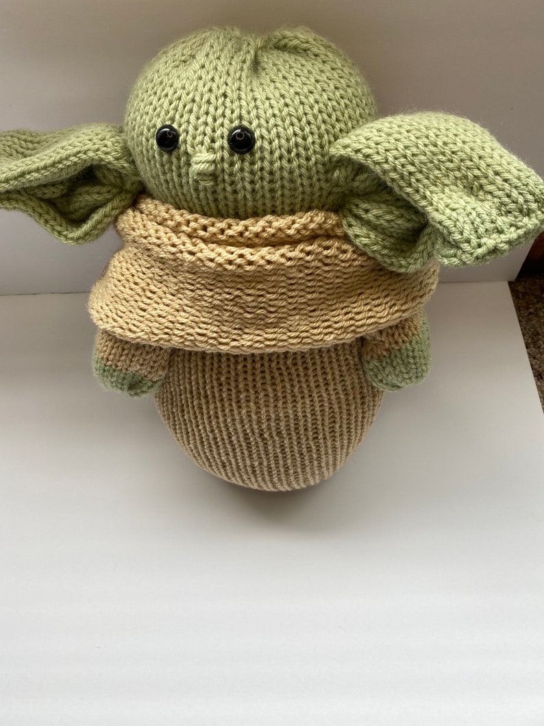May include: A knitted green and tan plush toy resembling a popular fictional character. The toy has large ears and a scarf.