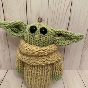 May include: A knitted green and tan plush toy with large black eyes. The toy is wearing a tan scarf.
