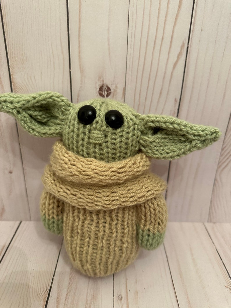 May include: A knitted green and tan plush toy with large black eyes. The toy is wearing a tan scarf.