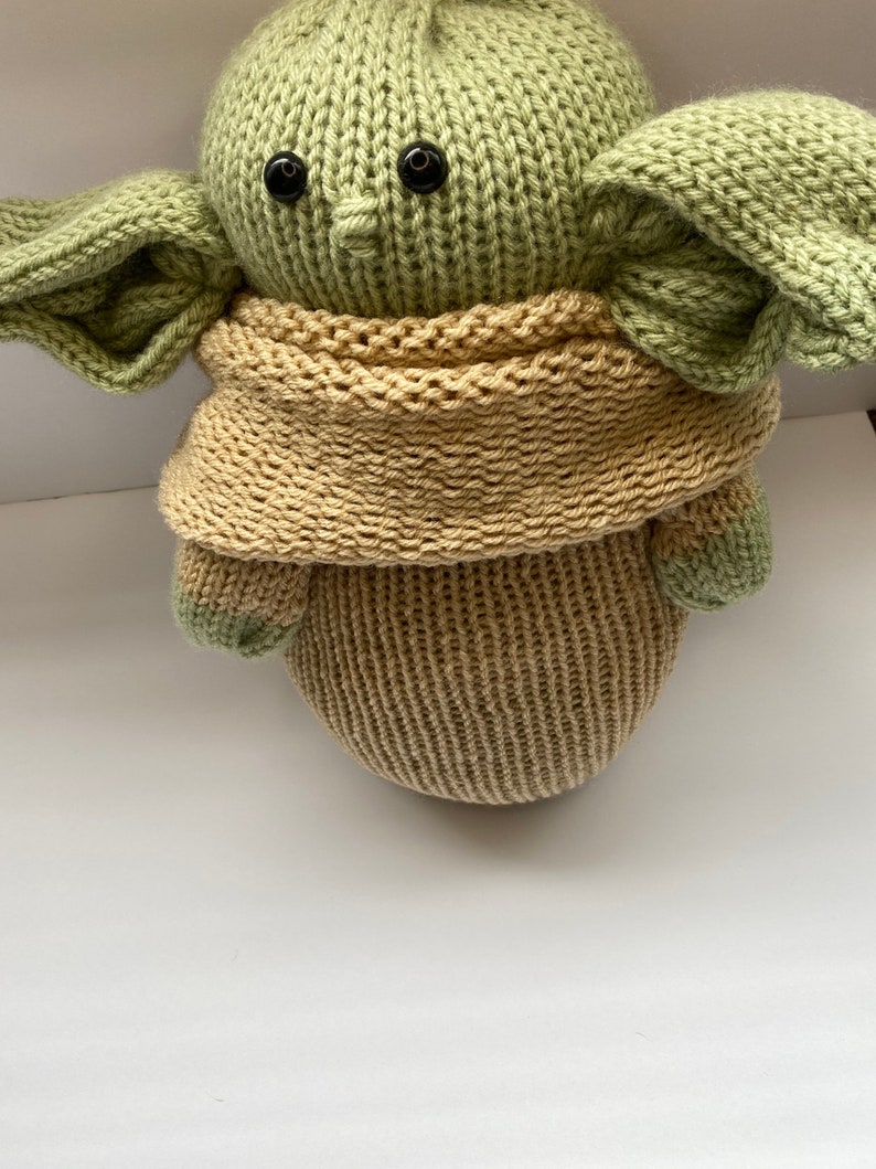 May include: A knitted green and brown plush toy with large ears and black eyes. The toy is wearing a brown knitted scarf.