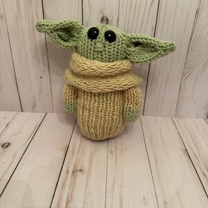 May include: A knitted green and tan plush toy resembling a character from the Star Wars franchise. The toy has large eyes and a small mouth.