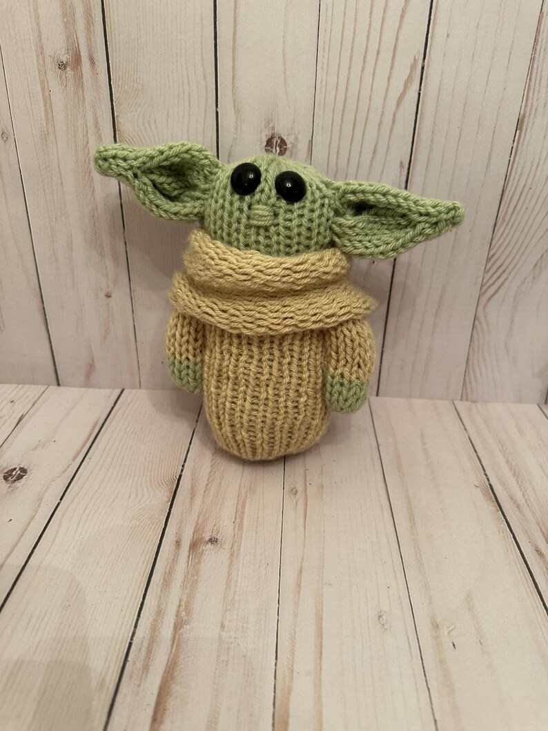 May include: A knitted green and tan plush toy resembling a character from the Star Wars franchise. The toy has large eyes and a small mouth.