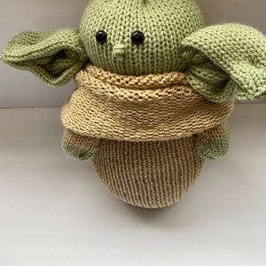 May include: A green knitted plush toy with large ears and black button eyes. The toy is wearing a tan knitted scarf.