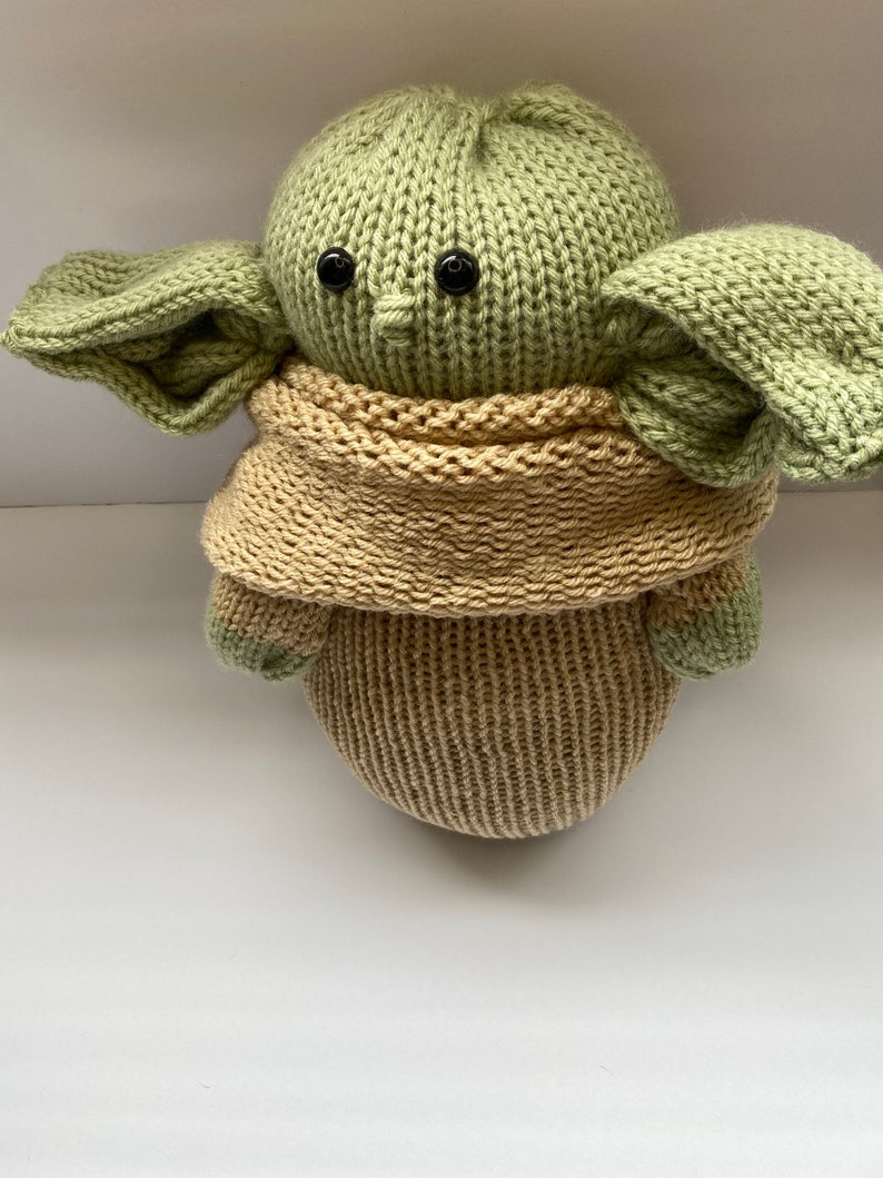 May include: A green knitted plush toy with large ears and black button eyes. The toy is wearing a tan knitted scarf.