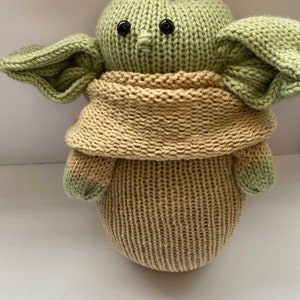 May include: A knitted green and tan plush toy resembling a popular fictional character. The toy has large ears, a small nose, and black button eyes.