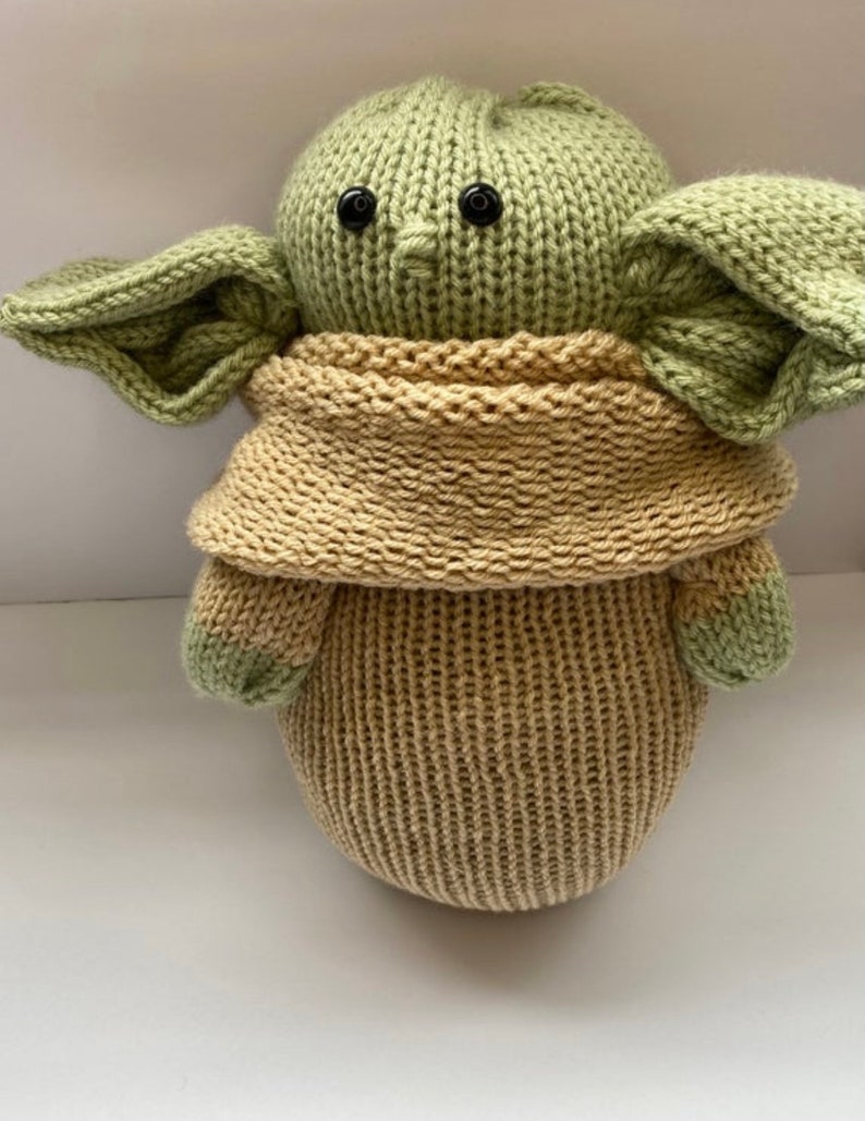 May include: A knitted green and tan plush toy resembling a popular fictional character. The toy has large ears, a small nose, and black button eyes.