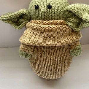 May include: A knitted green and tan plush toy resembling a character from the Star Wars franchise. The toy has large ears, a small nose, and black button eyes.