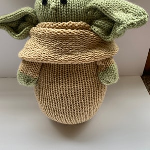 May include: A knitted green and tan plush toy resembling a character from the Star Wars franchise. The toy has large ears, a small nose, and black eyes.