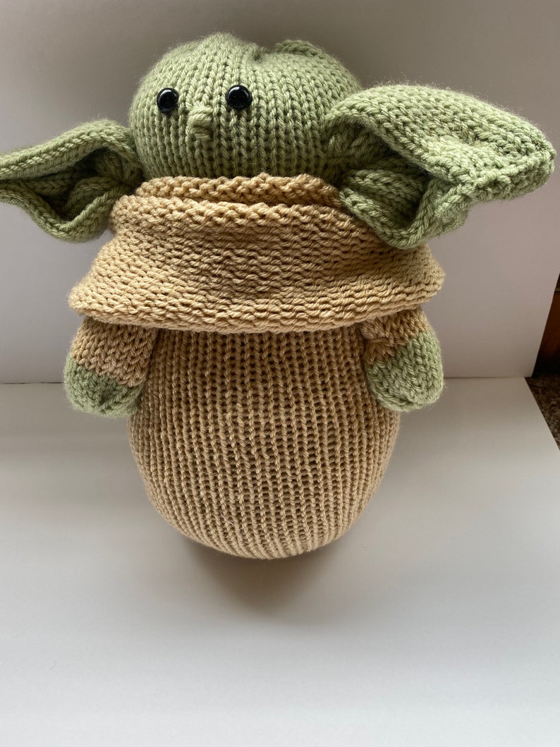 May include: A knitted green and tan plush toy resembling a character from the Star Wars franchise. The toy has large ears, a small nose, and black eyes.