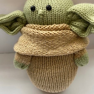 May include: A knitted green and brown plush toy resembling a character from the Star Wars franchise. The toy has large ears, a small nose, and black button eyes.