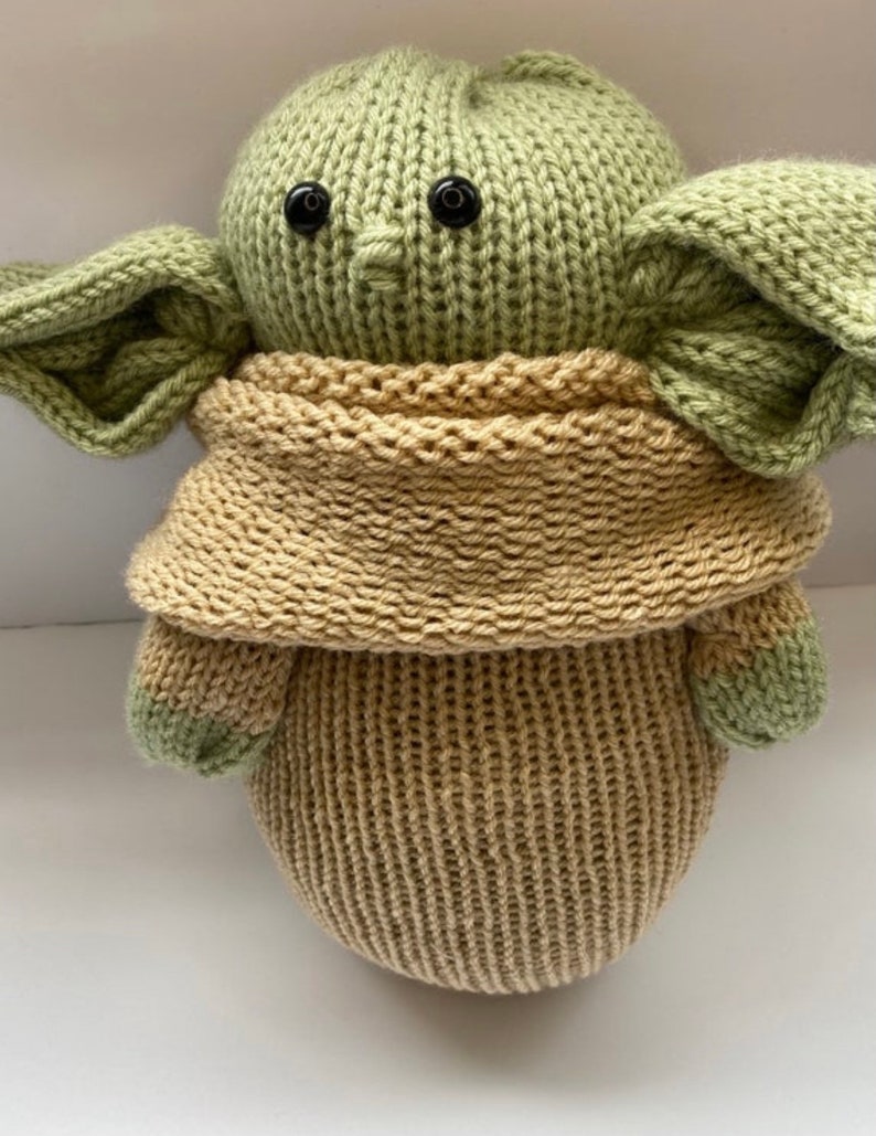 May include: A knitted green and brown plush toy resembling a character from the Star Wars franchise. The toy has large ears, a small nose, and black button eyes.
