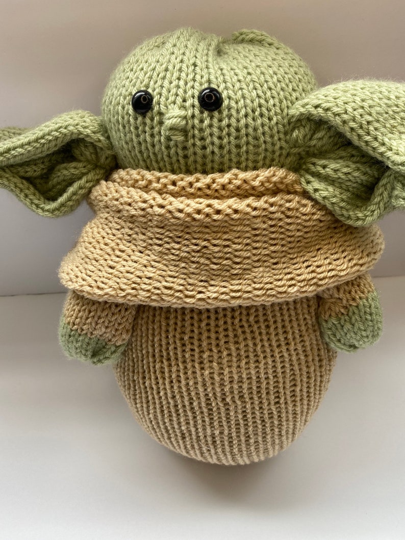 May include: A knitted green and tan plush toy with large ears and black eyes. The toy is wearing a tan knitted scarf.