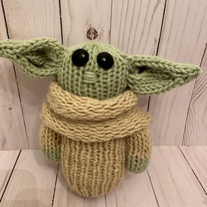 May include: A knitted green and beige plush toy with large black eyes. The toy is wearing a beige knitted scarf.