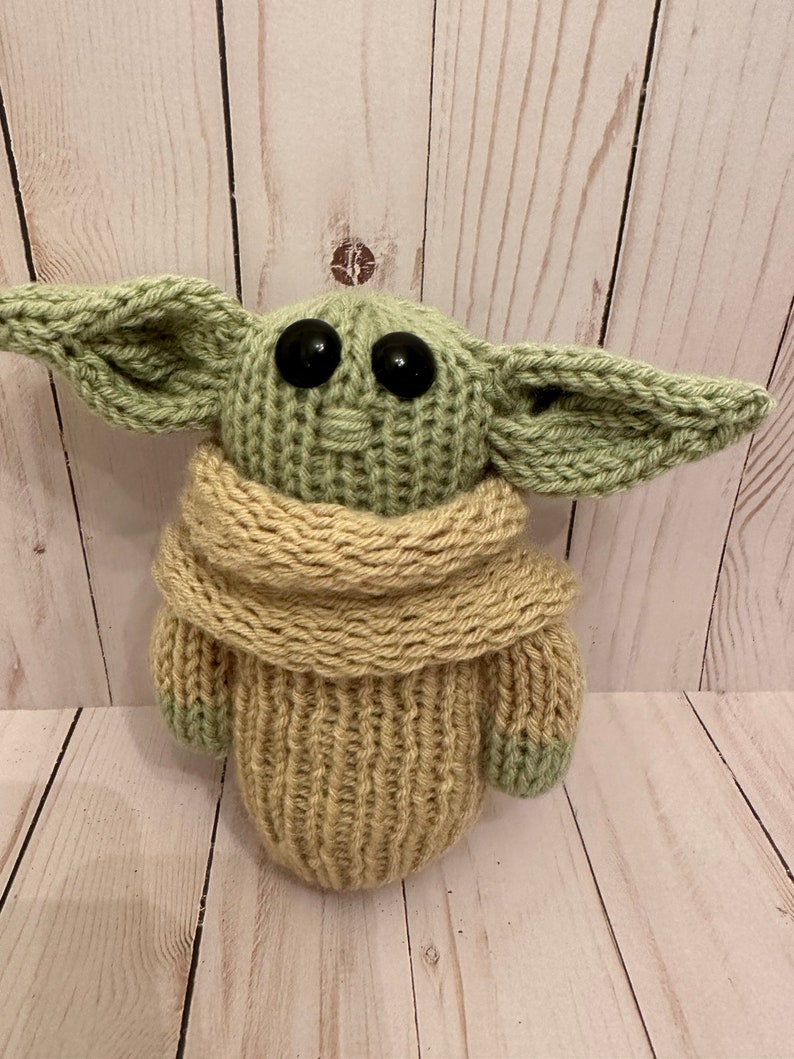 May include: A knitted green and beige plush toy with large black eyes. The toy is wearing a beige knitted scarf.