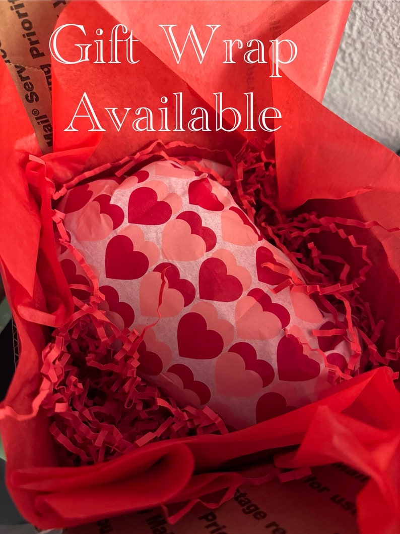 May include: A gift wrapped in red tissue paper and red crinkle paper, inside a cardboard box. The gift is wrapped in white paper with red and pink hearts. The text "Gift Wrap Available" is written in white script.