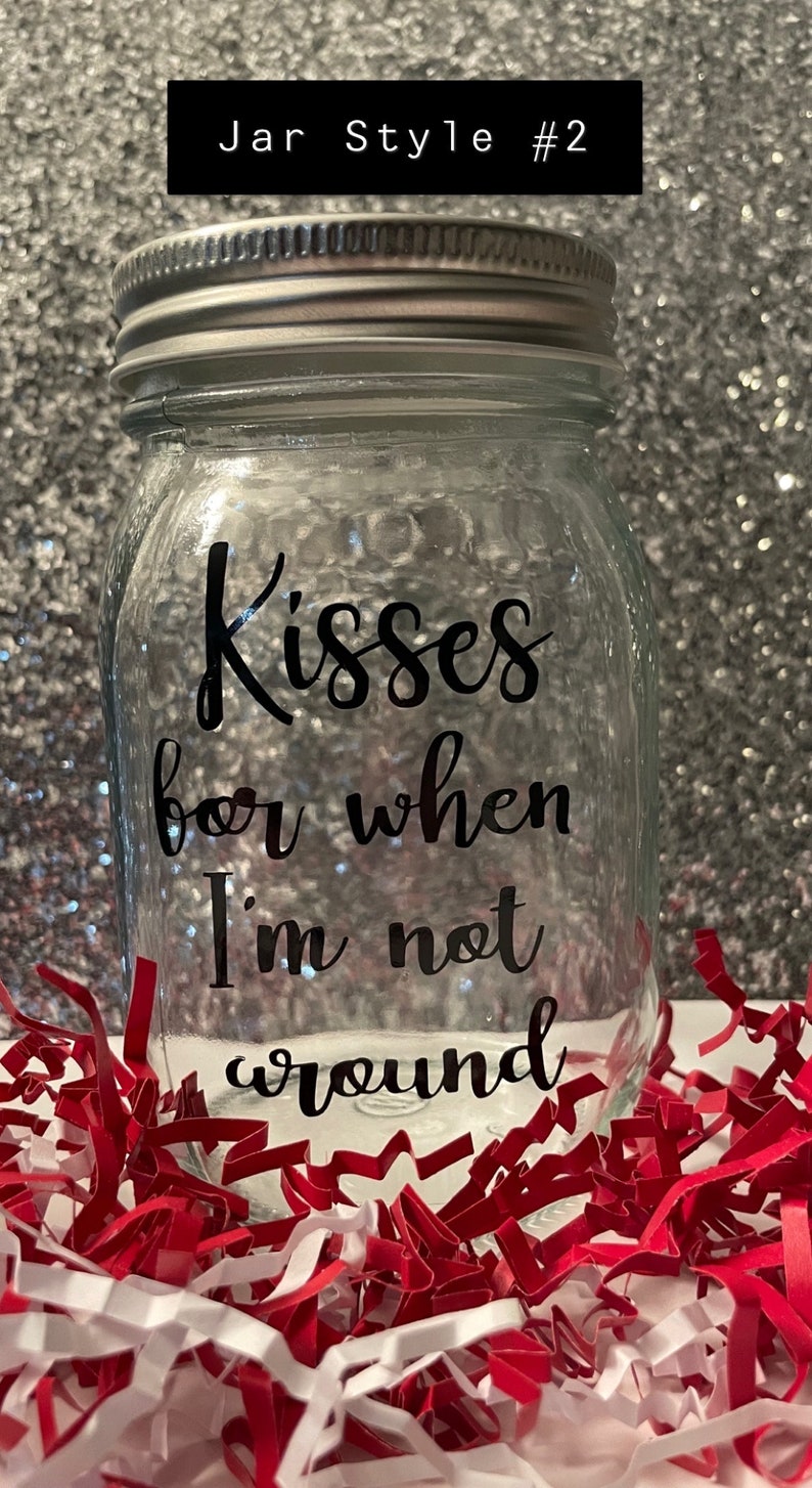May include: Clear glass jar with a silver lid, inscribed with the words "Kisses for when I'm not around" in black script. The jar is surrounded by red and white paper shreds. The text "Jar Style #2" is in a black rectangle.