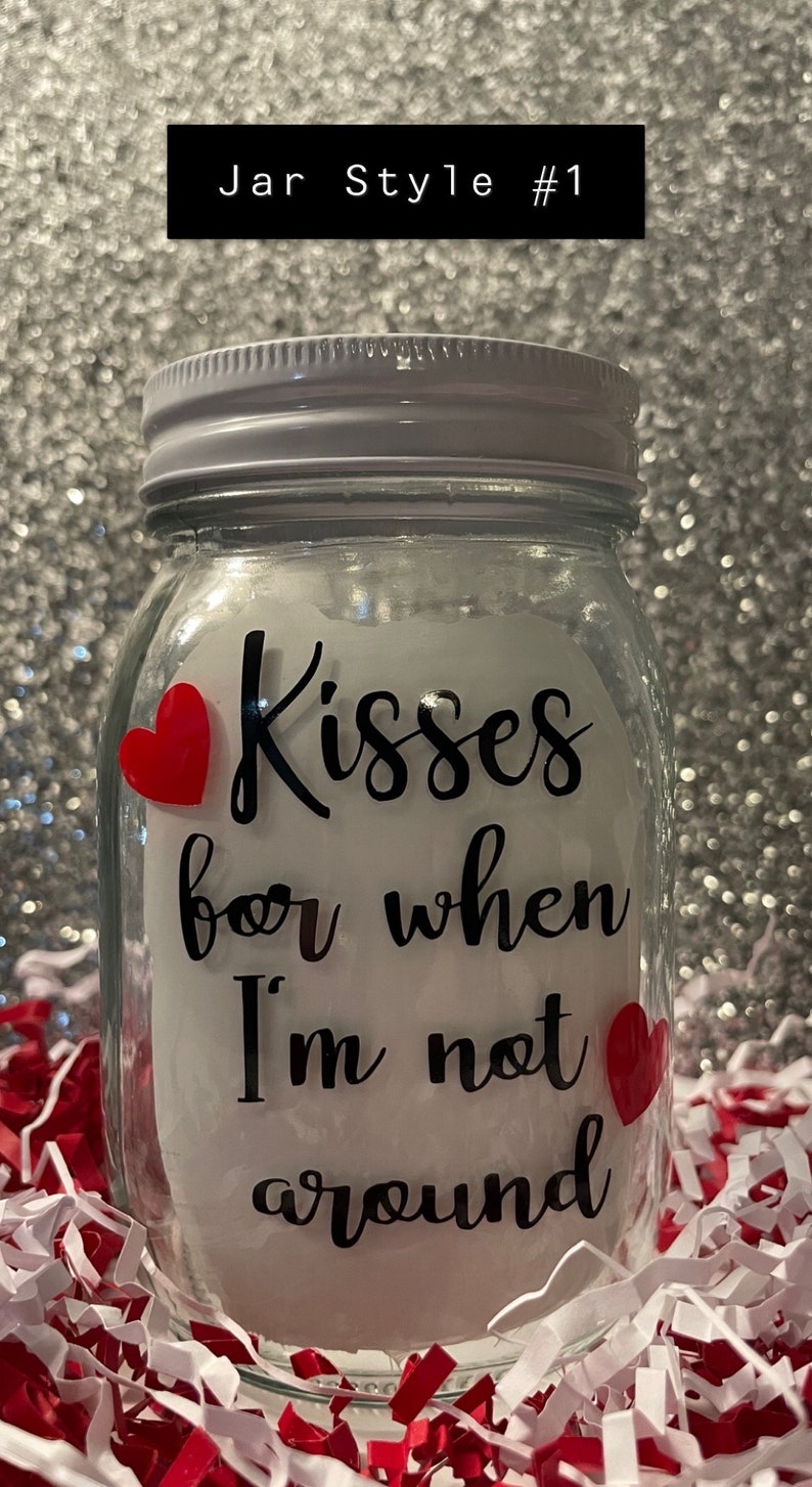 May include: A clear glass jar with a white lid and the text "Kisses for when I'm not around" in black script. Two red heart stickers are on the jar. The jar is surrounded by red and white paper shreds.